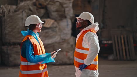 A-brunette-girl-in-a-blue-uniform-and-an-orange-vest-communicates-with-a-man-with-Black-skin-in-a-white-uniform-while-they-are-at-a-large-waste-processing-and-sorting-plant