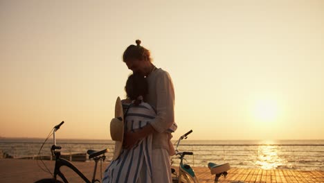 Glückliches-Paar,-Mann-Und-Frau,-Umarmen-Sich-Am-Strand-In-Der-Nähe-Des-Meeres-Bei-Sonnenaufgang-Im-Sommer.-Ein-Mann-In-Leichter-Kleidung-Und-Ein-Mädchen-In-Einem-Weißen-Und-Blauen-Hemd-Kamen-Mit-Fahrrädern-Am-Strand-An