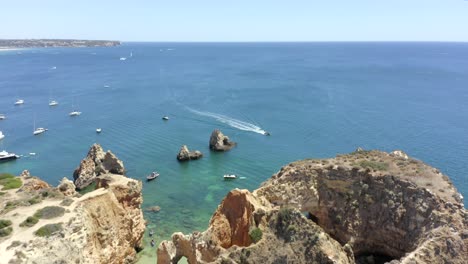 Drone-flies-over-Epic-Oceanside-rock-formations-before-finding-a-powerboat-speeding-across-the-blue-sea,-Aerial