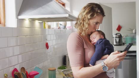 video de una madre caucásica con un bebé recién nacido en un portero usando un teléfono inteligente en la cocina