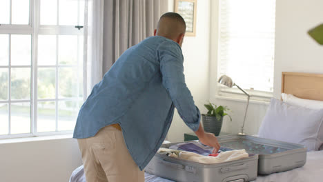 Happy-biracial-man-packing-suitcase-in-bright-bedroom