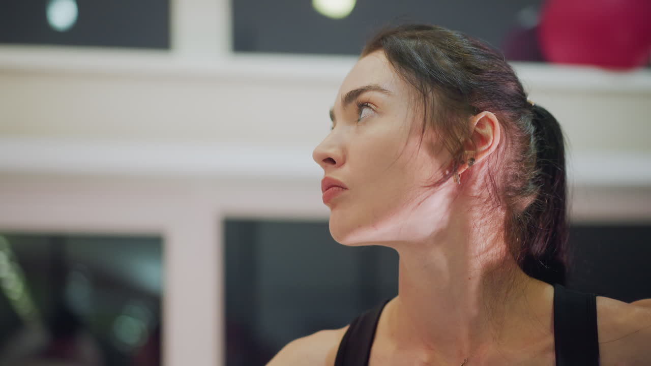 donna bianca di profilo che si allena in studio, postura da allenatrice durante una ripetizione controllata, linea del collo evidenziata da luce di contorno, coda di cavallo che oscilla con il movimento, riflessi della finestra e palla rosa