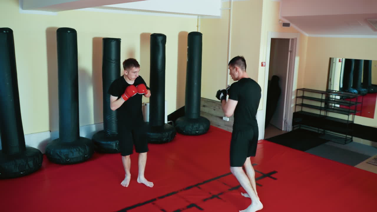 Fighters facing off inside martial arts gym, wearing gloves and black sportswear, preparing for sparring on red mat with focus, balance, and defensive stance during combat training session