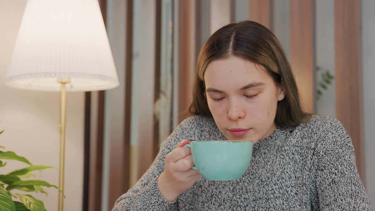 Young adult enjoying aroma of hot beverage with calm satisfaction holding cup in both hands cozy indoor setting soft lamp light nearby phone on table background decor includes potted plant