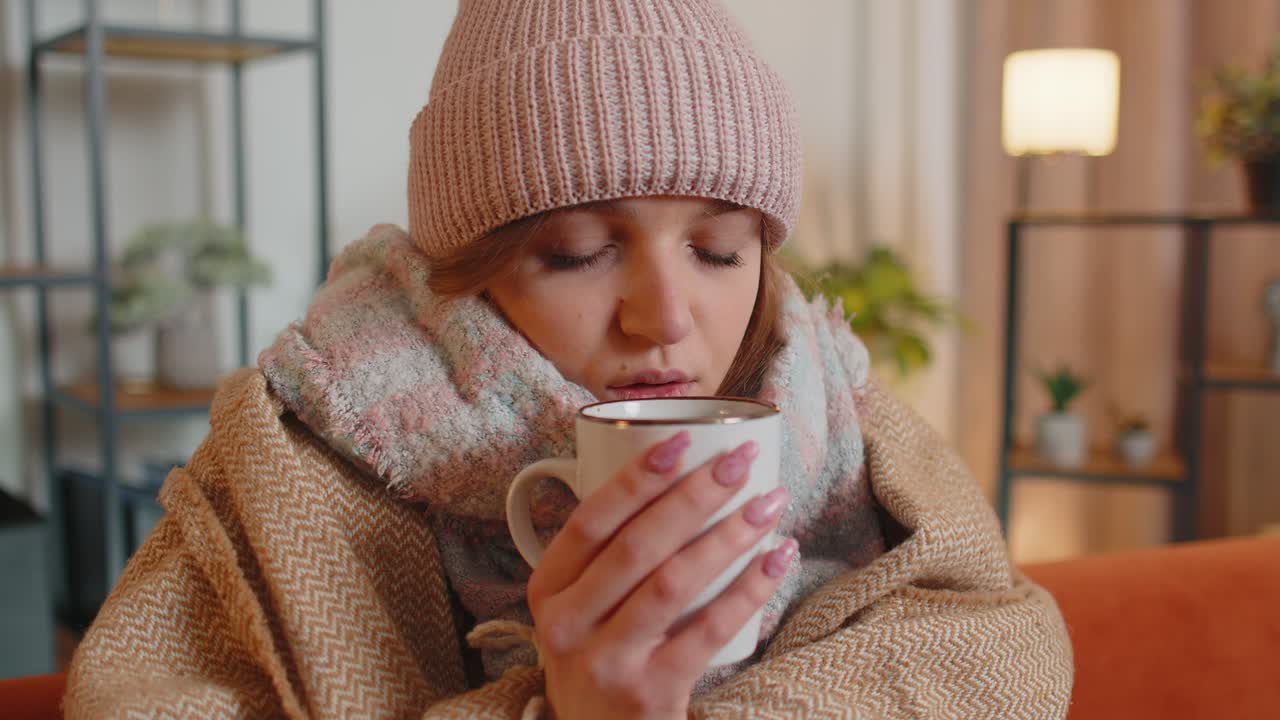 병든 젊은 여성은 모자를 입고, 카레드에 싸여, 혼자 앉아서, 소파에서 추위에서 떨며, 뜨거운 차를 마시고 있다.