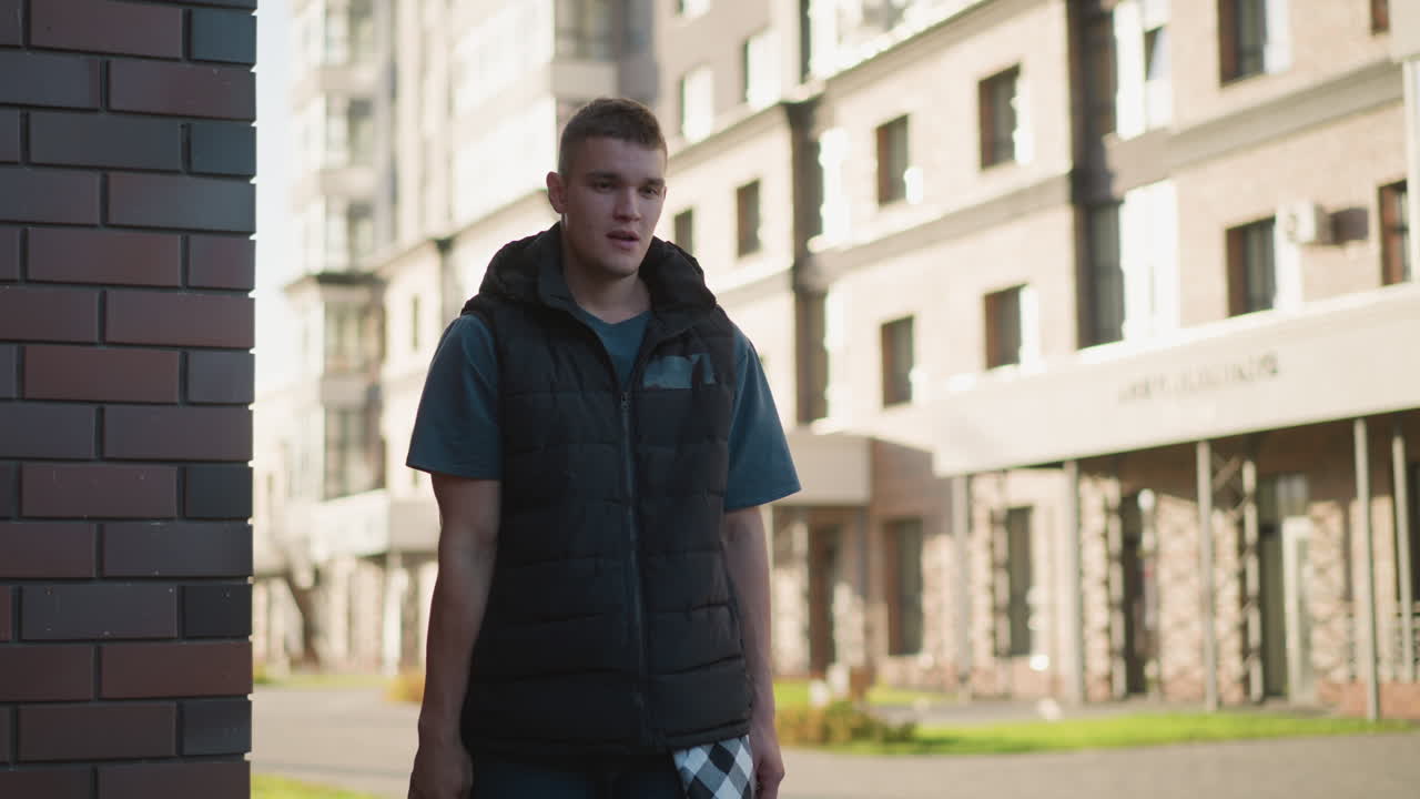 young man smiles lightly while tossing cigarette in relaxed gesture near urban building with soft sunlight casting shadows on brick wall and modern city architecture in blurred background