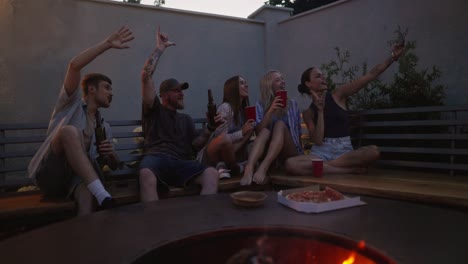 A-happy-and-cheerful-group-of-friends-takes-a-selfie-while-sitting-on-a-bench-near-a-barbecue-with-a-fire-during-their-party-in-the-evening-in-the-backyard-of-a-country-house