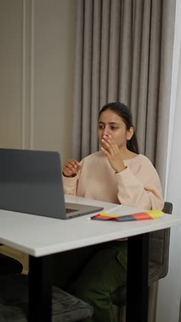 Vertical-video-of-a-happy-brunette-girl-in-a-beige-sweater-sitting-in-front-of-a-gray-laptop-and-communicating-via-video-with-a-teacher-while-studying-a-foreign-language-German-in-a-modern-apartment-during-the-day