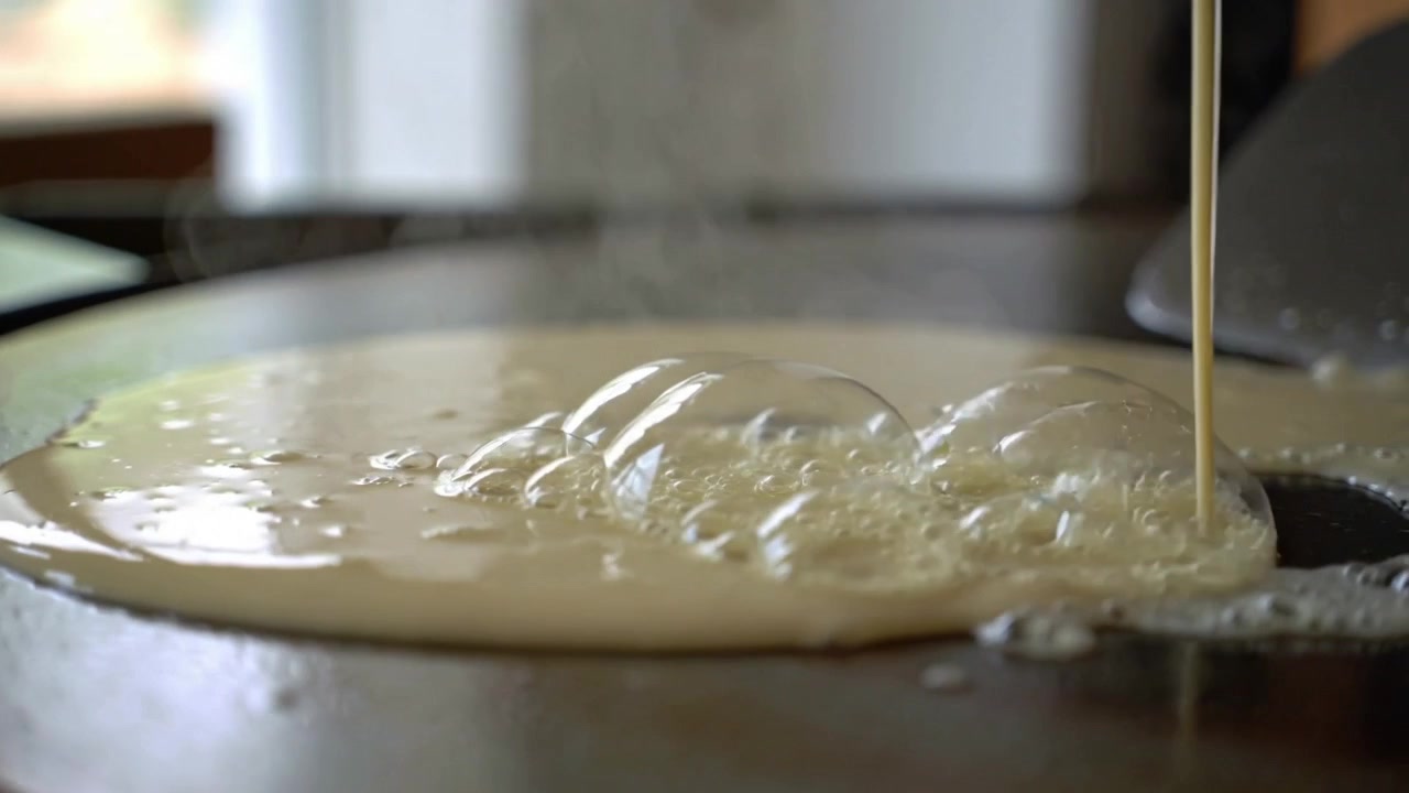 Pouring batter stream forming crepe disk on griddle to cook, making bubbles, steam, spatula