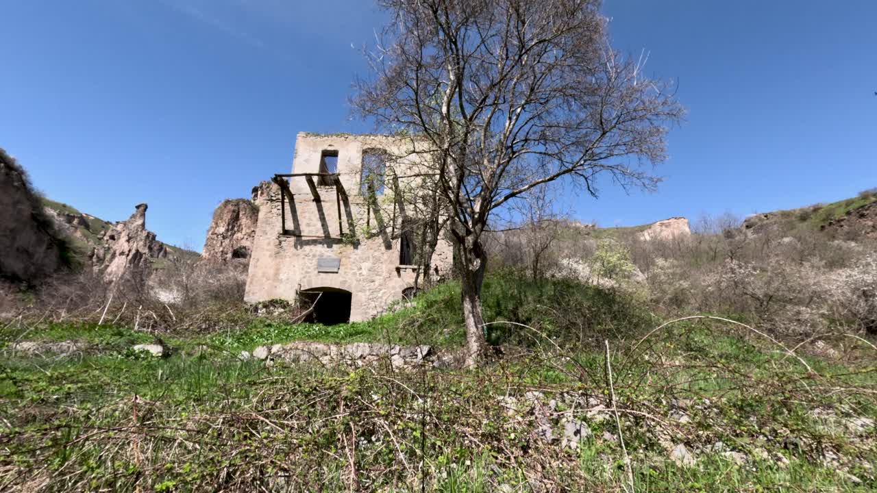 Un vecchio edificio si erge in una valle circondato da piante verdi e alberi. Il sole splende luminoso e la natura cresce tutt'intorno. L'area è pacifica e piena di vita