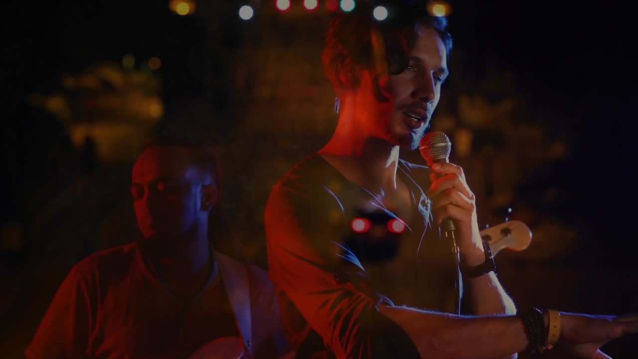 Singer adjusting mic grip then singing for music while bassist plucking, lights drifting over stage