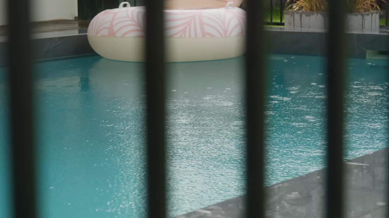 Privat swimmingpool set gennem mørkt poolhegn, der viser kraftig regn på overfladen og omgivelserne. Ubrugt poollegetøj i baggrunden.