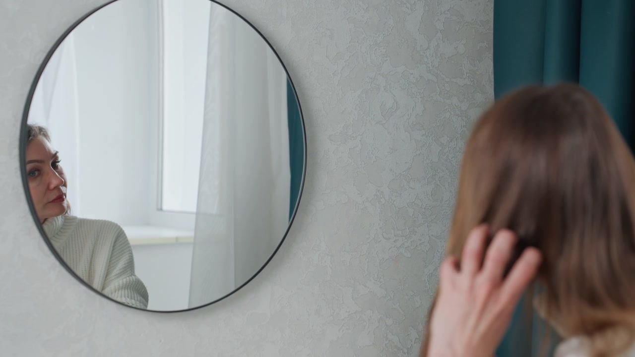 Coach donna caucasica che prova le battute davanti a uno specchio rotondo, indossando un maglione di lana vicino a una tenda color verde acqua, sistemando i capelli e la postura mentre studia il suo riflesso, preparazione concentrata per una performance.