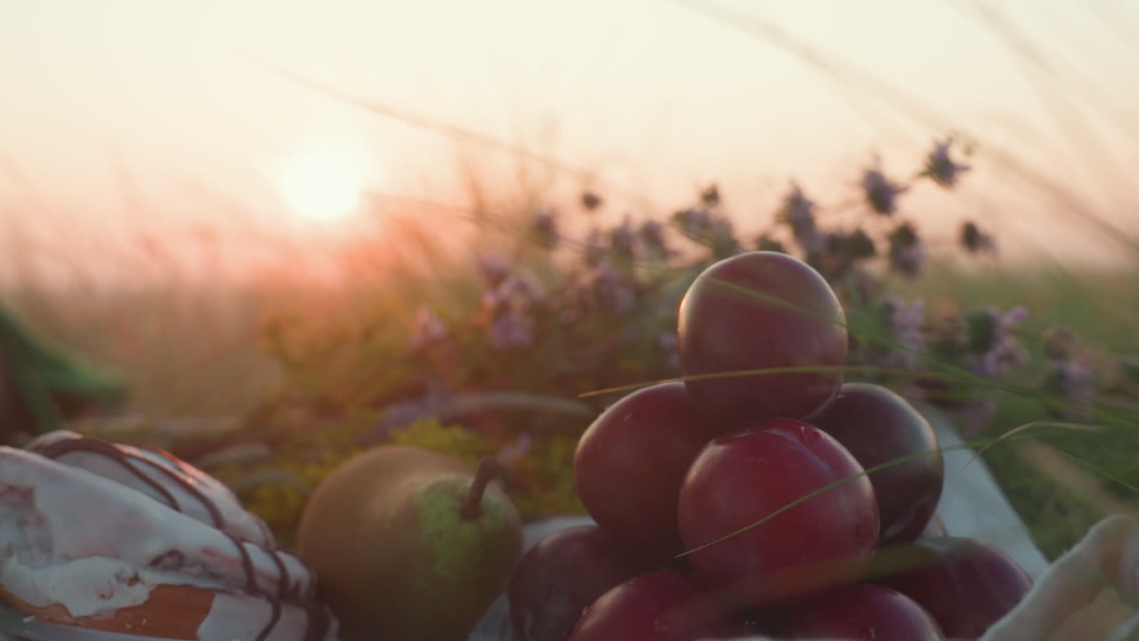 Close up of hand reaching to pick ripe plum from stacked fresh fruit plate during golden hour sunset picnic in tall grassy field with pastries, pear, wildflowers, dew, kissed petals