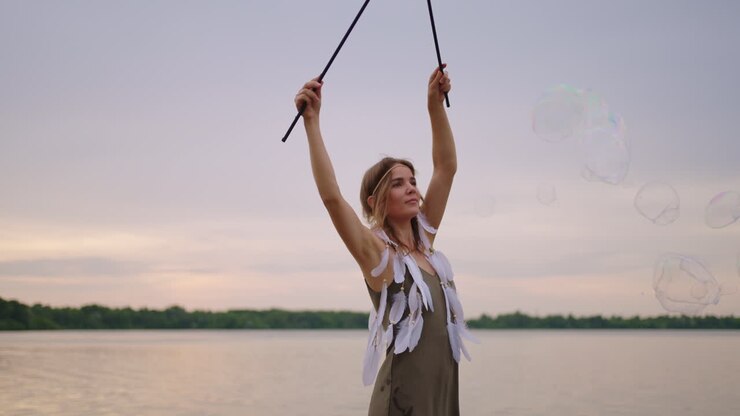 Una giovane donna hippie in abito e con piume in testa crea enormi bolle di sapone al tramonto sulla riva di un lago, al rallentatore.