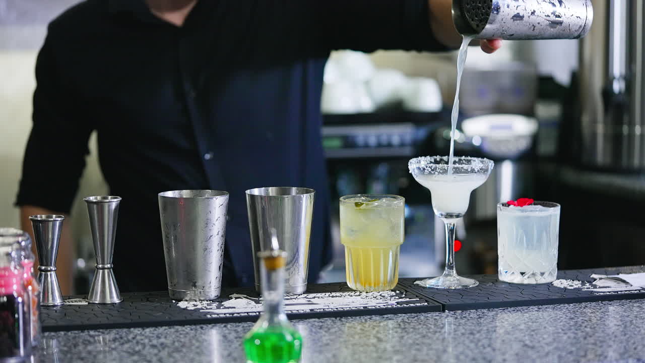 Barman in black shirt filling the wineglass with beverage. Glasses for cocktails making at the bar stand. Close up. Blurred backdrop.