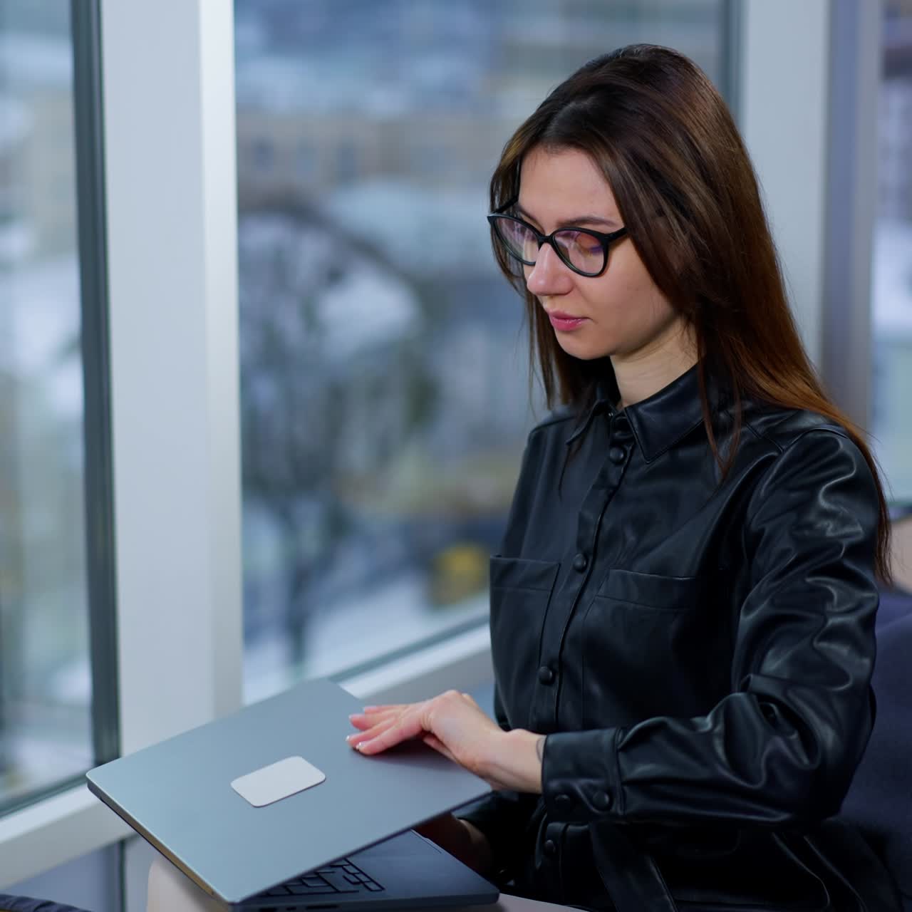 Long-haired lady wearing black jacket sits in bean bag chair near the window. Business woman takes a laptop from a little coffee table to start work
