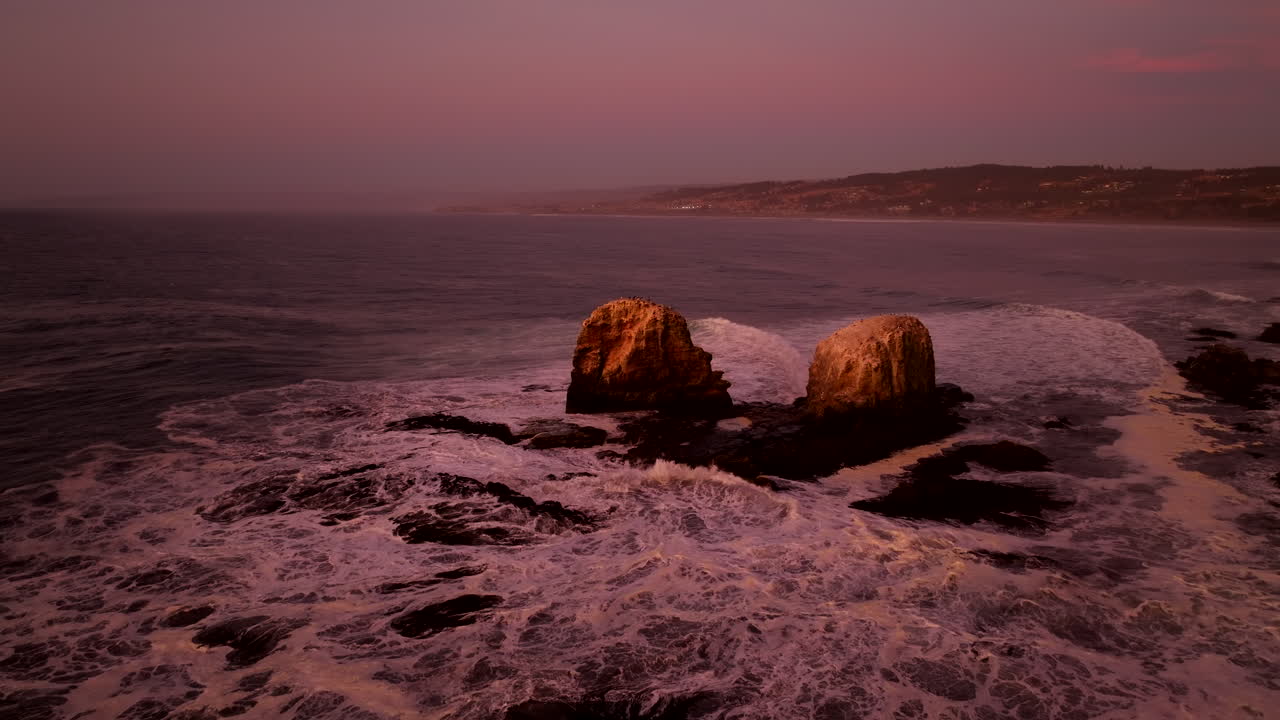 Sunset drone push in toward boulders with pelicans on top on Chilean coastline