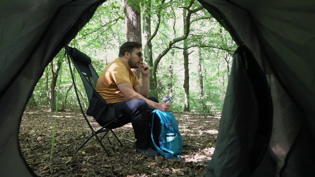 Mand spiser og hviler siddende på en stol på en campingplads i skoven.