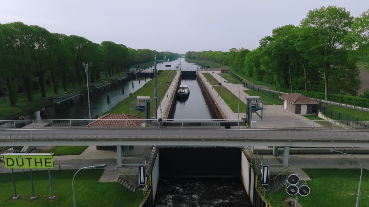 Downward aerial movement showing a barge navigating into Doerpen Lock, surrounded by forested canal banks under soft daylight.