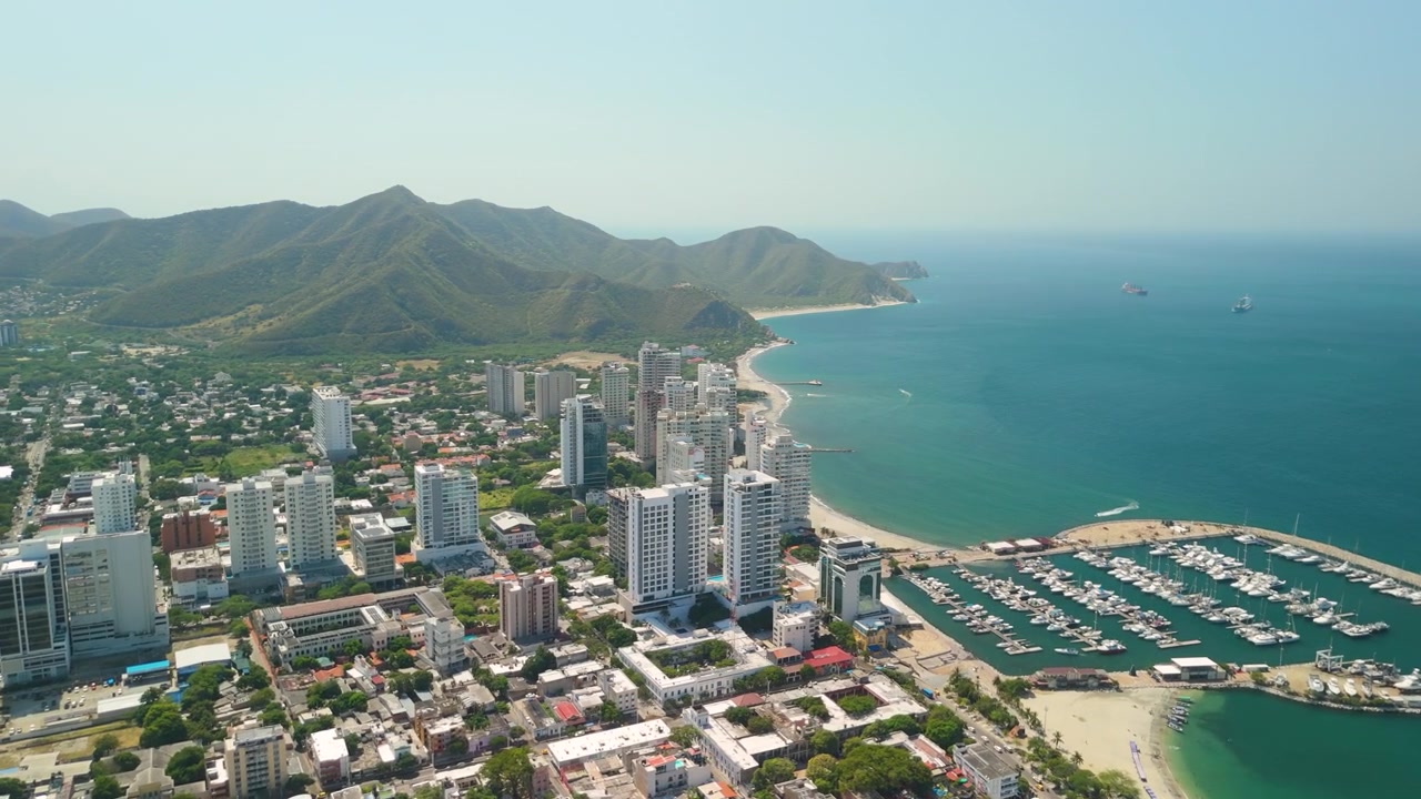 Parallax aéreo de alta altitude ao redor da Baía de Santa Marta com iates da Marina Internacional, praia, horizonte costeiro e as montanhas da Sierra Nevada na Colômbia