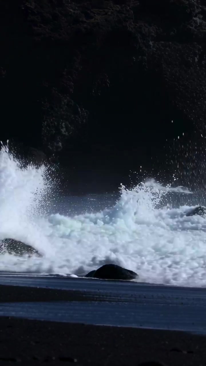 Vertical video: Wind pushing large ocean wave crashing on black sand, spraying foam over sea stack