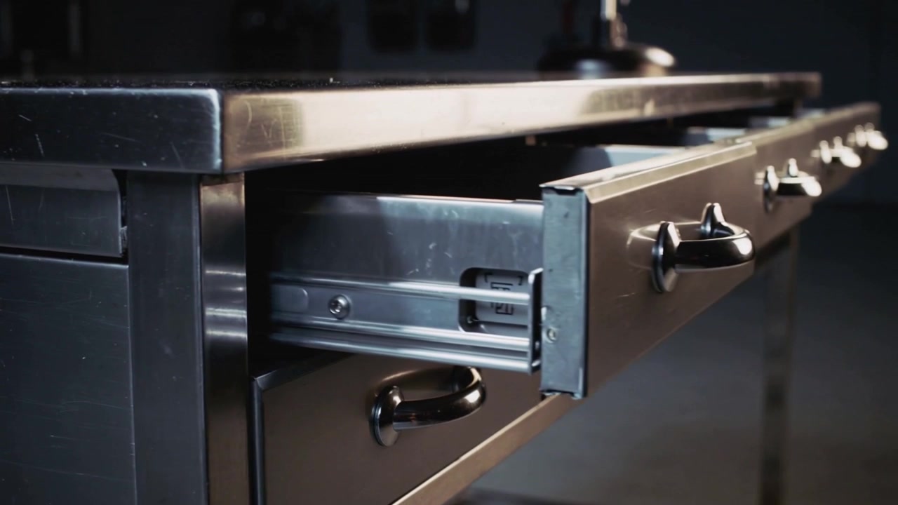 Sliding steel bench drawer after handle pull showing telescopic runner in dim shop, exposing slides