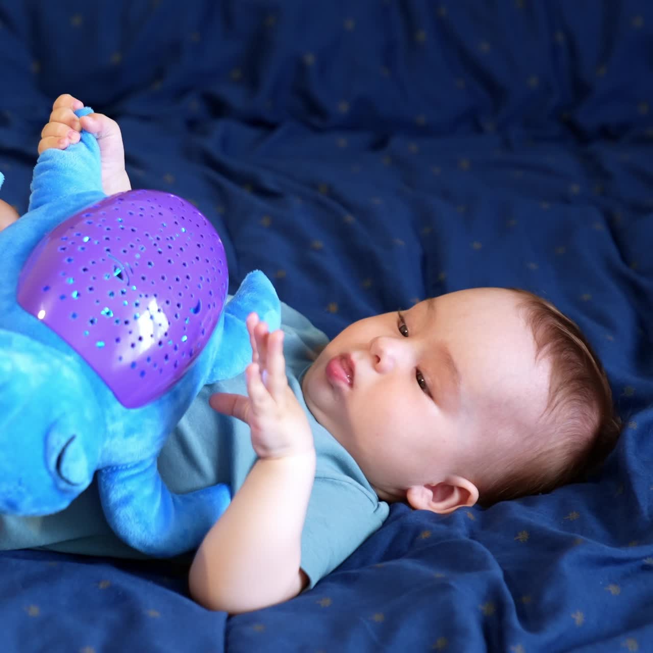 Cute Caucasian toddler lies on the soft bed on his back. Baby boy holding a big toy turtle on his belly playing with it. Side view