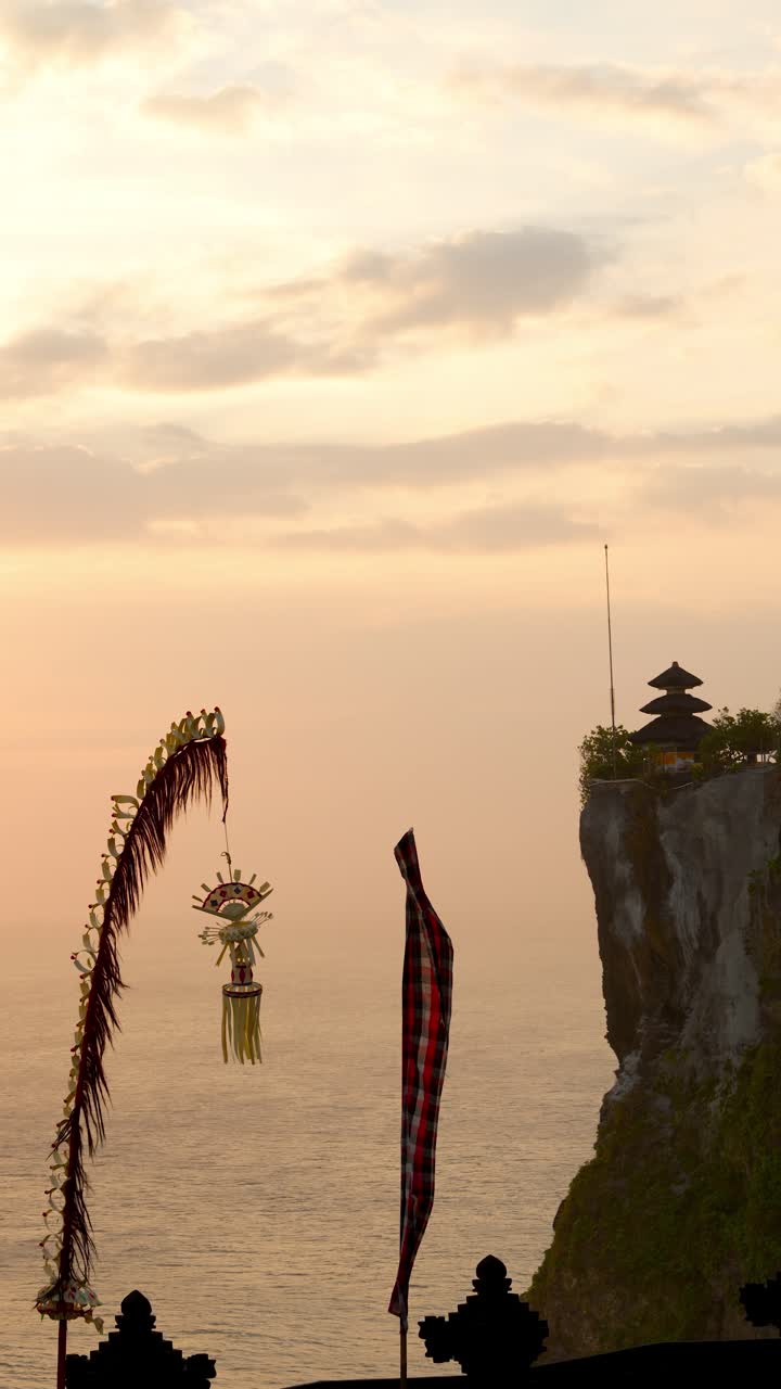 Golden hour view of Uluwatu Temple on a limestone cliff above the Indian Ocean; penjor and ceremonial flags sway in the breeze. Serene Bali seascape, vertical handheld static