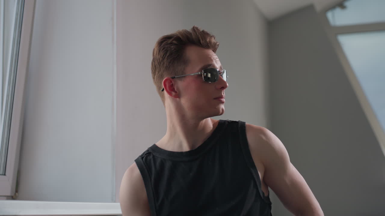 Teenager with geometric neck tattoo seated by large tilted window in modern loft inspects dark sunglasses before sliding them on, soft natural light highlighting his profile, muscles, and urban style
