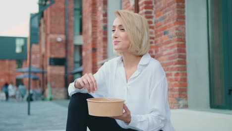 attractive blond businesswoman in a white shirt eating a salad on a city street. healthy lunch blonde girl. healthy lifestyle and eating concept