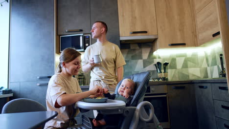 mother feeds her lovely baby boy sitting at high chair. smiling man with a mug in hand stands behind looking at his family.
