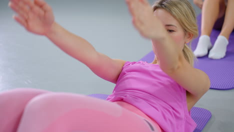 mujer con traje de entrenamiento rosa realiza ejercicios de sentada, involucrando los músculos del núcleo mientras se acuesta en una alfombra de yoga púrpura, vista parcial de las piernas de otra persona en el fondo
