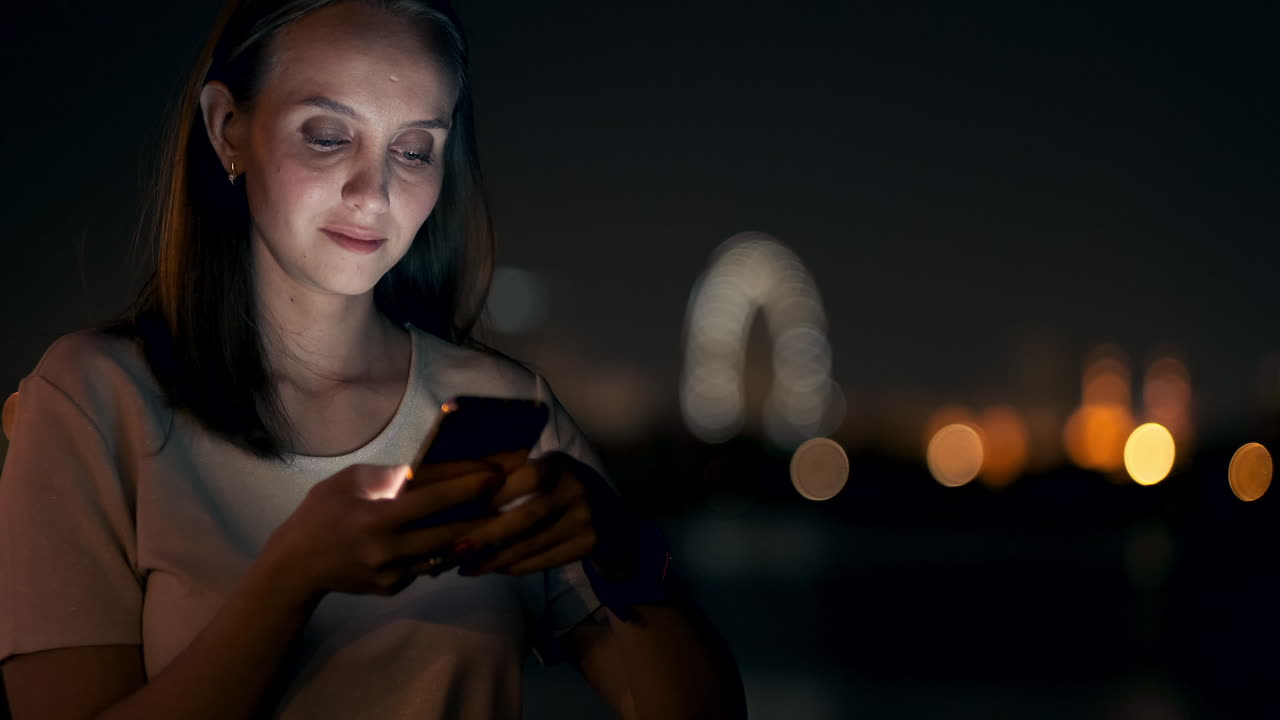 A young woman looks into the smartphone and writes text messages on the Internet against the backdrop of the night city. Girl businessman working on vacation remote work via mobile phone. Gadget addiction