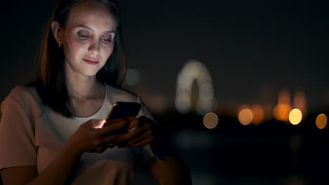 a young woman looks into the smartphone and writes text messages on the internet against the backdrop of the night city. girl businessman working on vacation remote work via mobile phone. gadget addiction