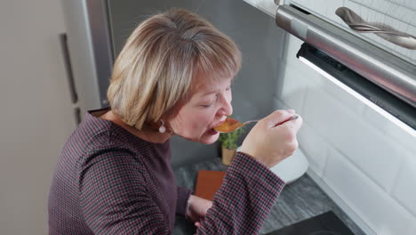 vista aérea de una mujer joven probando comida recién preparada bajo una brillante luz de cocina, ella sostiene una cuchara de sopa, saboreando el sabor, con un libro de cocina descansando en la encimera