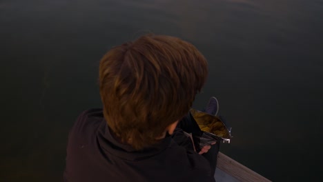 vista de arriba hacia abajo de un joven sentado en el borde de un muelle sobre un lago comiendo de una bolsa de papas fritas