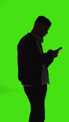 Silueta-De-Vídeo-Vertical-De-Un-Hombre-Con-Auriculares-Inalámbricos-Enviando-Mensajes-De-Texto-En-Un-Teléfono-Móvil-Contra-Una-Pantalla-Verde