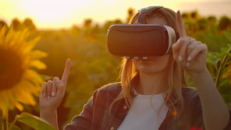 la adolescente con el cabello largo en camisa a cuadros y vaqueros está trabajando en gafas vr. ella está involucrada en el proceso de trabajo. es una agradable noche soleada en el campo de girasoles al atardecer.