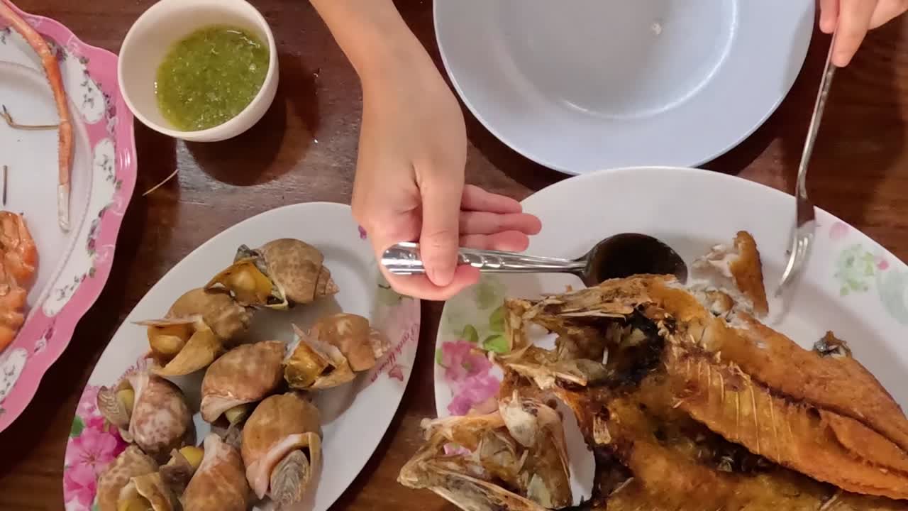 Close-up of hands using utensils to serve fried fish and shellfish with green sauce on a wooden table.