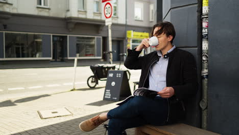 a young man enjoys his coffee break outdoors while reading a magazine. dressed in casual-business attire, he relaxes in an urban environment with traffic passing in the background.