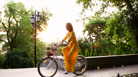 Schöne-Frau-In-Langem-Gelben-Kleid,-Die-Im-Morgengrauen-Ein-Stadtfahrrad-Mit-Einem-Korb-Und-Blumen-Darin-Fährt.-Linseneffekt