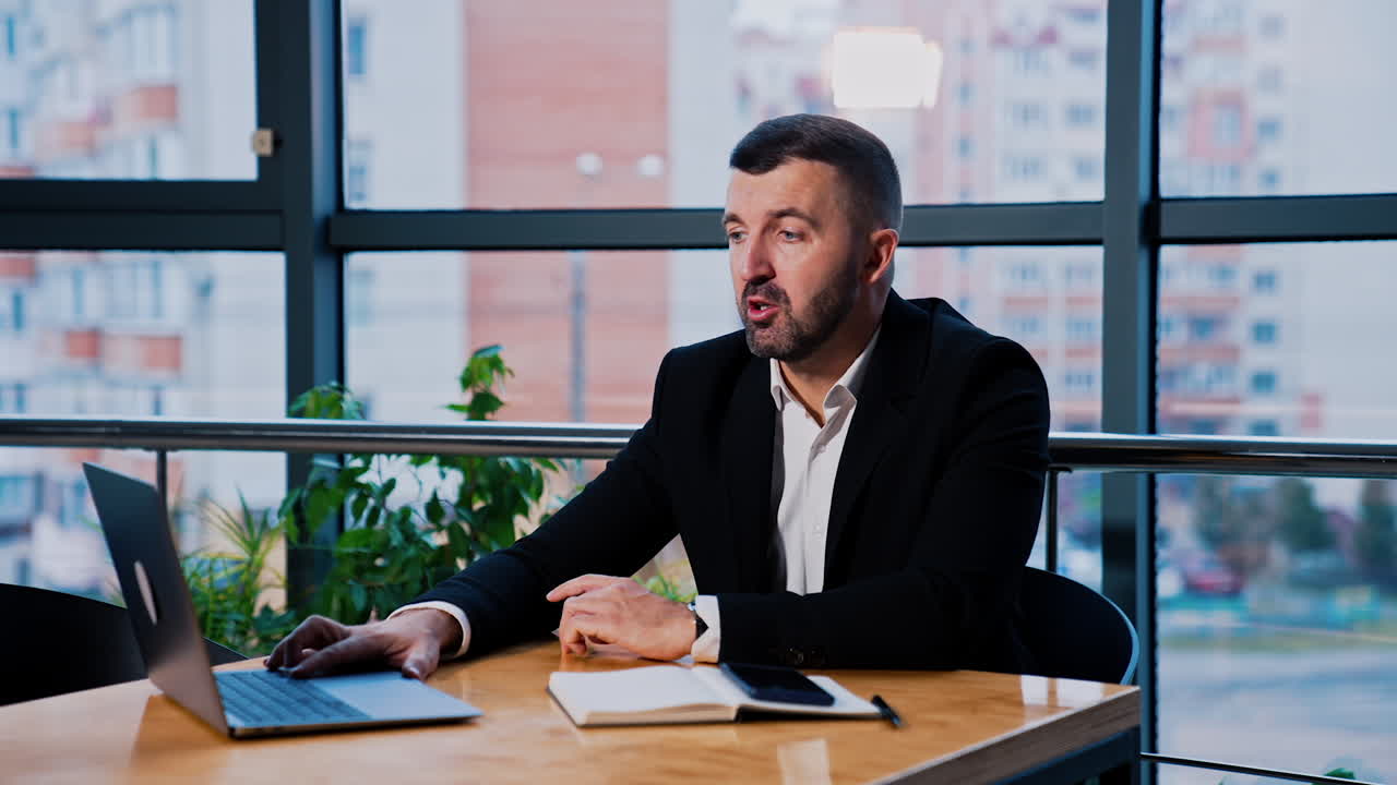 Adult mid-aged man sitting in the office at the desk. Bearded man working at laptop in front of him. Businessman having online chat with partners.
