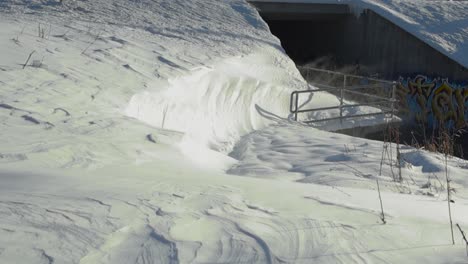 Left to right panning video of white snow being blown and gushed by wind during winter sunny day in slow motion. The wind is moving the white fluffy snow under a bridge, over a railing, by a river.