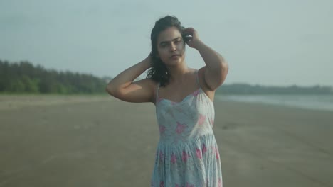 A-woman-stands-on-a-sandy-beach,-adjusting-her-hair-as-the-breeze-blows-gently