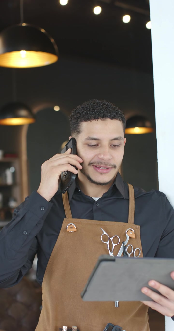 Video verticale: barbiere afroamericano con grembiule da lavoro, al telefono, che usa il tablet per confermare gli appuntamenti.