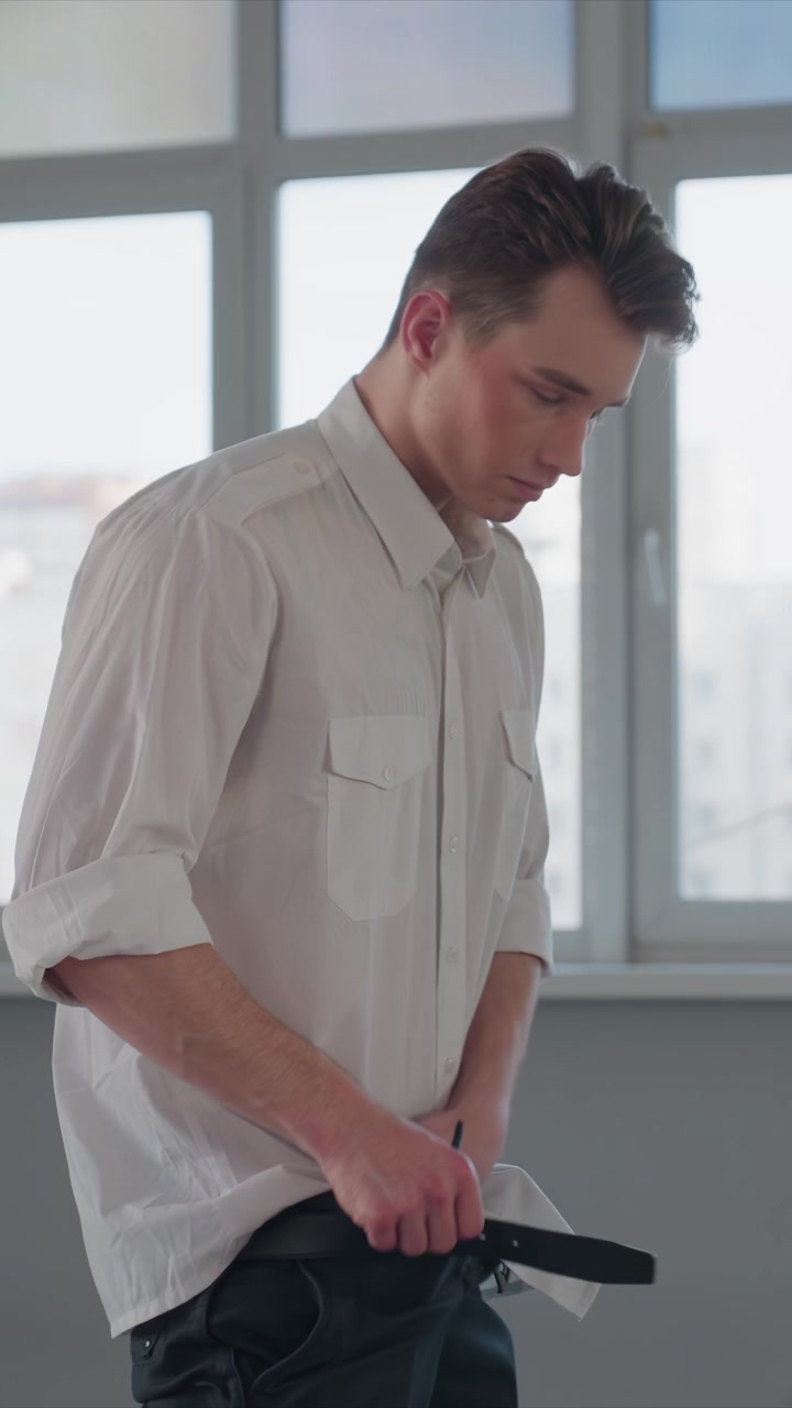 Giovane uomo in camicia bianca abbottonata si sistema i vestiti all'interno. L'uomo si arrotola le maniche e infila il tessuto nei pantaloni neri. La scena è luminosa con la luce del giorno che filtra attraverso i vetri delle finestre.