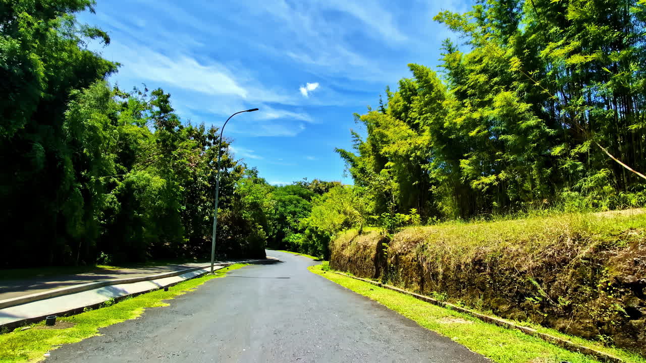 A beautiful rear-mounted point-of-view shot from a moving car captures the scenic, empty road receding through a lush green bamboo forest on the way to the GWK Cultural Park in Bali