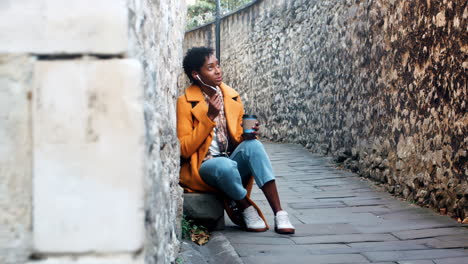 Young-woman-wearing-a-yellow-pea-coat-and-blue-jeans-sitting-in-an-alleyway-in-a-historical-city-talking-on-her-smartphone-using-earphones,-full-length,-selective-focus