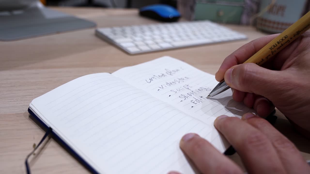 Close-up of a person writing a to-do list in a lined notebook on a wooden desk, with keyboard and computer in soft focus, creating a productive home office workspace scene